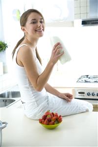 Mujer, sentada en la mesa de la cocina, con fresones y una botella de leche en la mano.