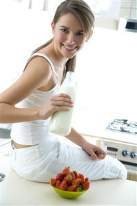 Mujer, sentada en la mesa de la cocina, con fresones y una botella de leche en la mano.