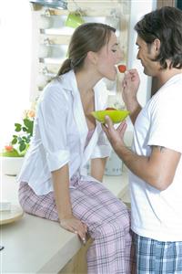 Pareja en la cocina. El, le pone un freson en la boca.