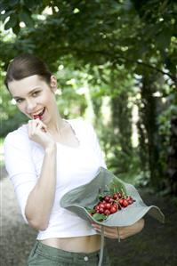 Jardineria. Mujer cogiendo una cereza de un sombrero lleno de cerezas recien cogidas. 