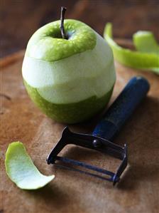 Half-peeled green apple with peeler on wooden background