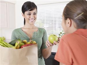 Woman handing young girl in kitchen an apple out of grocery bag and smiling