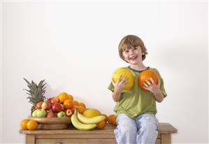 Young boy sitting on kitchen island with assorted fruit holding pumpkins and smiling