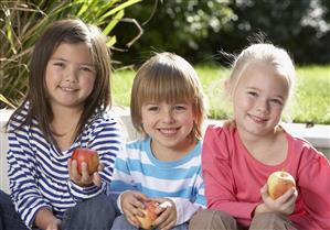 Three young kids sitting outdoors holding apples and smiling