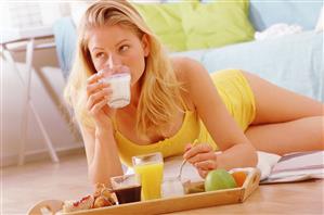 Mujer desayunando tumbada en el suelo. Delante tiene una bandeja con el desayuno. Ella toma un vaso de leche.