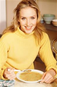 Mujer comiendo sopa con verduras. Lleva un jersey amarillo de cuello alto.