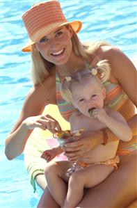 Mujer sentada al borde de la piscina con su hija pequeña, comiendo macedonia de frutas.