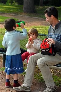 Padre sentado en un banco del parque mientras sus hijas meriendan.